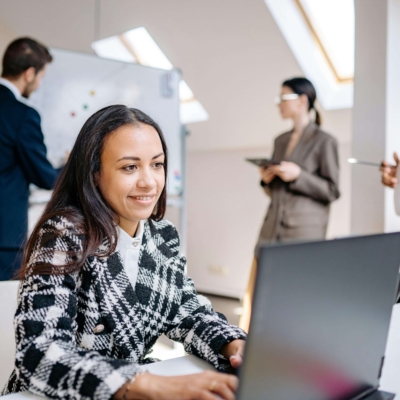 Woman wearing a smart blazer at a laptop with a meeting happening behind her.
